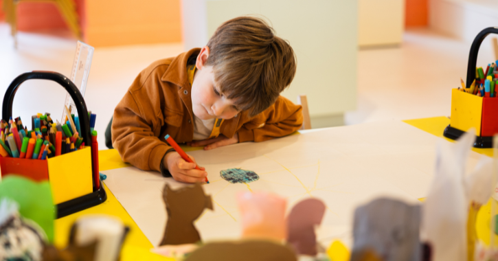 Photograph of a young child focused on drawing at a table in a brightly lit classroom or art space. Table holds colourful pens and pencils in yellow holders, with various art supplies and paper cutouts visible in foreground and background.